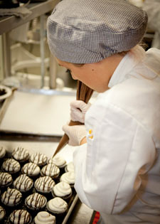 A student pastry chef decorating a batch of freshly baked small cakes.